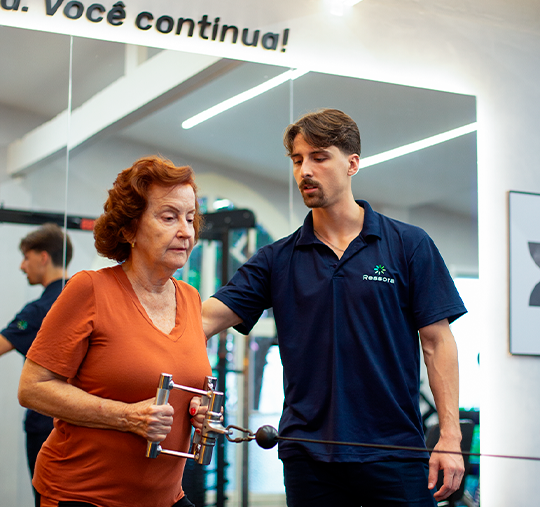 Mulher idosa com cabelos ruivos realiza exercício de remada em aparelho de cabos, acompanhada de perto por um fisioterapeuta da clínica Ressora, em ambiente de reabilitação.